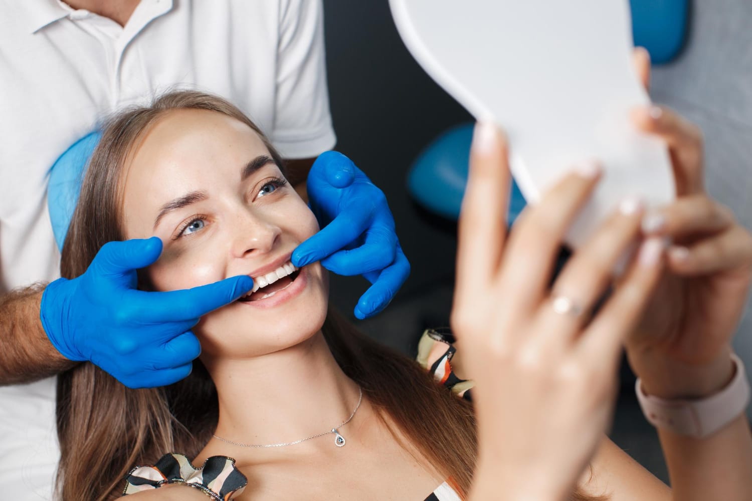 Woman smiling and looking in a mirror while a dentist in blue gloves examines her teeth.