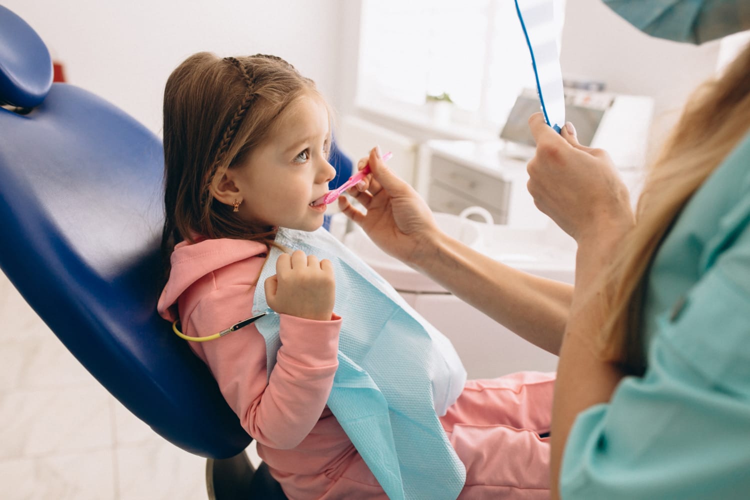 A young girl sitting in a dental chair while a dentist brushes her teeth with a pink toothbrush.
