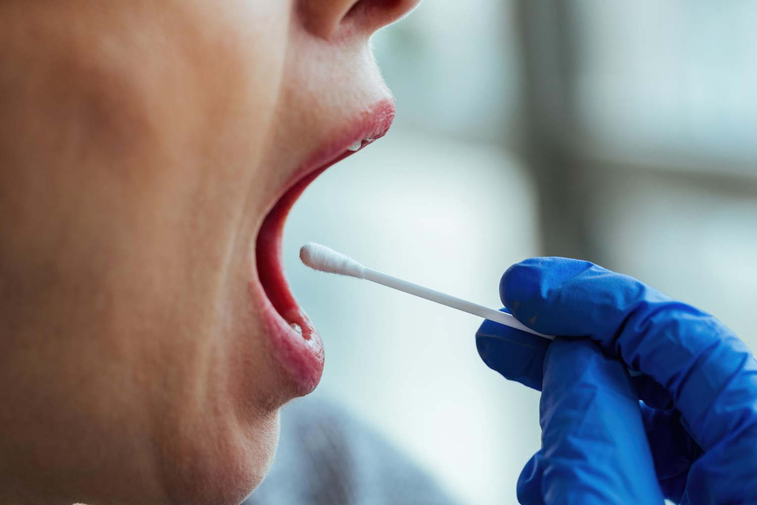 A gloved hand uses a cotton swab to take an oral sample from a person’s open mouth.